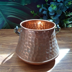 May include: A copper-colored hammered metal pot with two small handles. The pot has a wide, flared shape and a textured surface. The interior of the pot is also copper-colored. The pot is sitting on a wooden surface.