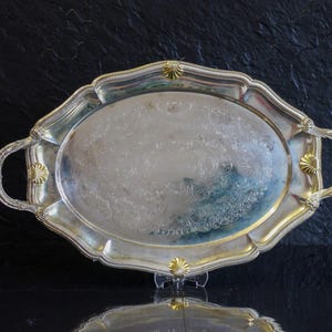 Può includere: Vassoio ovale argentato con manici ornati e bordo decorativo. Il vassoio presenta un motivo floreale dettagliato al centro e dettagli dorati. Il vassoio è esposto su una superficie nera.