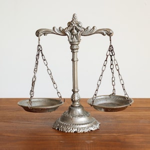 May include: A silver-coloured decorative balance scale with two round trays suspended by chains. The scale has an ornate design and sits on a wooden surface. The background is a plain white wall.