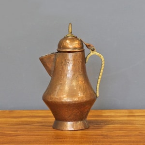 May include: A copper-colored pitcher with a lid and a brass handle. The pitcher has a textured surface and a spout. The handle is curved and has a decorative design. The pitcher is likely used for serving beverages.