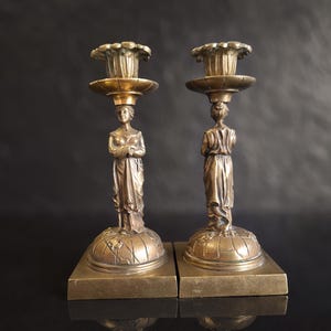 May include: Two antique brass candlesticks. Each candlestick features a classical female figure supporting a floral-shaped candle cup. The figures stand on a globe base atop a square pedestal. The candlesticks are displayed against a dark background.