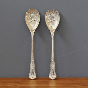 Könnte beinhalten: Ein silbernes Servierlöffel- und Gabel-Set. Der Löffel und die Gabel haben verzierte florale Designs auf den Köpfen und Griffen. Das Set wird auf einer Holzoberfläche vor grauem Hintergrund präsentiert.