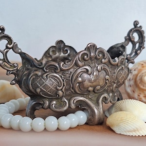 Op de afbeelding: Een sierlijke zilverkleurige kom met krullende handvatten en decoratieve details. Een witte kralenarmband en schelpen zijn bij de kom geplaatst. De kom staat op een houten oppervlak.