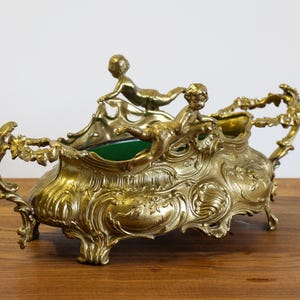 Peut inclure: Centre de table en métal doré orné de chérubins. Le récipient présente un intérieur vert et des poignées décoratives. Il est posé sur une surface en bois, mettant en valeur ses détails complexes et son esthétique antique.