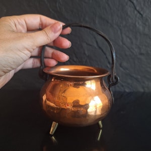 Puede incluir: Un pequeño caldero de cobre martillado de forma redonda con acabado metálico pulido, tres patas cortas de metal y un asa giratoria oscura fijada a los lados. El recipiente tiene una abertura ancha y un cuerpo redondeado.