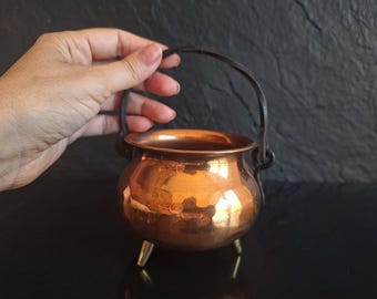 Puede incluir: Un pequeño caldero de cobre martillado de forma redonda con acabado metálico pulido, tres patas cortas de metal y un asa giratoria oscura fijada a los lados. El recipiente tiene una abertura ancha y un cuerpo redondeado.