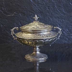 Può includere: Una zuppiera argentata con coperchio e dettagli ornati. Il piatto ha una base a piedistallo, un corpo arrotondato e due manici. Il coperchio è sormontato da un elemento decorativo. Lo sfondo è una superficie scura e strutturata.
