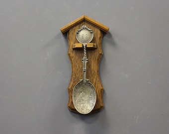 May include: A decorative spoon mounted on a wooden plaque. The spoon is silver-colored with a detailed design on the bowl and a twisted handle. The plaque is brown with a house-shaped top and a carved design. The spoon has the year 1981.