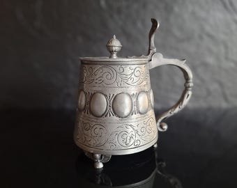 Puede incluir: Una jarra de peltre con tapa que presenta un patrón de volutas florales en relieve en las bandas superior e inferior, con una fila central de medallones ovalados. El recipiente incluye una tapa con bisagra, un asa curva y tres patas.