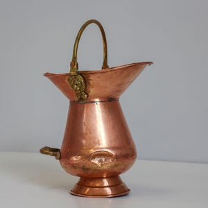 Puede incluir: Una jarra de metal color cobre con asa y boquilla de latón. La jarra tiene una parte superior acampanada y un cuerpo bulboso, con un adorno decorativo de latón cerca del asa. La jarra es probablemente vintage y adecuada para uso decorativo.