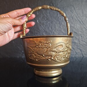 Pode incluir: Um balde de latão com alça oscilante apresentando um design em relevo de um dragão entre nuvens. O recipiente de metal tem uma base circular e uma abertura larga, exibindo um acabamento dourado metálico com detalhes em relevo no exterior.