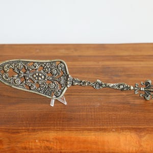 Puede incluir: Un servidor de pastel antiguo de color plateado con un diseño calado ornamentado. El servidor presenta un detallado patrón floral y de volutas en la hoja y el mango, con una flor de lis al final del mango. El servidor se exhibe sobre un soporte transparente.