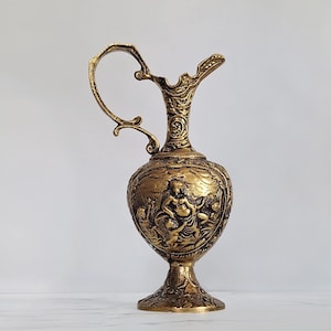 Könnte beinhalten: Verzierter goldfarbener Krug mit detailliertem Griff und Ausguss. Der Körper des Kruges weist ein Reliefdesign von Figuren auf. Der Krug steht auf einem Sockel, der eine antike oder Vintage-Ästhetik zeigt.