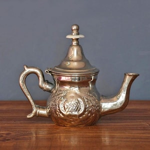 Op de afbeelding: Een zilverkleurige theepot met een gebogen handvat en tuit. De theepot heeft een decoratief bloemmotief en een deksel met een kleine, witte knop. Hij staat op een houten oppervlak tegen een grijze achtergrond.