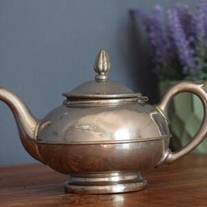 May include: A silver-colored teapot with a rounded body, a curved spout, and a loop handle. The lid is topped with a decorative finial. The teapot sits on a wooden surface, with a vase of purple flowers in the background.
