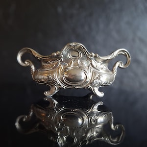Op de afbeelding: Een sierlijk zilverkleurig serveerschaaltje met krullende handvatten en decoratieve details. De schaal heeft een ovale centrale vorm en staat op kleine voetjes. Het reflecterende oppervlak eronder weerspiegelt het ontwerp van de schaal.