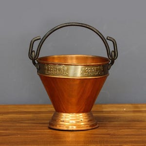 Puede incluir: Un cubo de cobre con un asa negra y una banda decorativa de latón. El cubo tiene forma cónica y una base redonda. La banda de latón presenta un diseño en relieve. El cubo se exhibe sobre una superficie de madera.