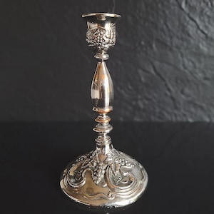 May include: A silver-toned metal candlestick holder featuring an embossed grape and vine motif on the circular base and the top candle cup. The stem is smooth and tapered, connecting to a wide, ornate base with detailed relief work.
