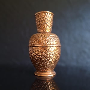 May include: A copper-colored decorative container with a geometric pattern. The container has a bulbous base, a mid-section with a lid, and a smaller, flared top. The surface is covered in a repeating triangular design, creating a textured appearance.