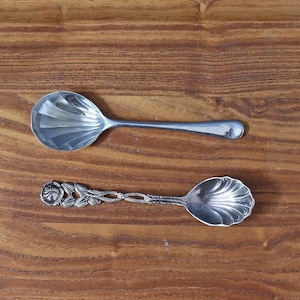 Può includere: Due cucchiai d'argento su una superficie di legno. Un cucchiaio ha una ciotola semplice e arrotondata e un lungo manico. L'altro cucchiaio ha un manico decorativo con dettagli floreali e di foglie e una ciotola smerlata. I cucchiai sono probabilmente per servire.