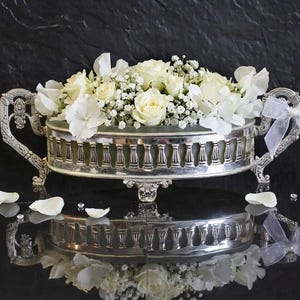 Puede incluir: Un centro de mesa plateado lleno de rosas blancas, hortensias y paniculata. El cuenco tiene asas ornamentadas y está decorado con una cinta blanca. Pétalos de flores blancas y pequeños cristales están esparcidos alrededor de la base.