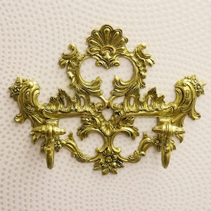 Könnte beinhalten: Verzierte goldfarbene Wandleuchte mit symmetrischem Design. Die Leuchte weist aufwendige Blumen- und Schnörkeldetails mit zwei Kerzenhaltern auf. Der Hintergrund ist eine strukturierte, cremefarbene Oberfläche.
