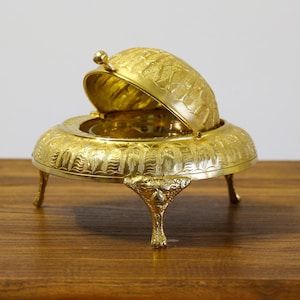 Puede incluir: Un plato de servir dorado y ornamentado con una tapa abovedada, apoyado sobre tres patas decorativas. La tapa está parcialmente abierta, revelando el interior. El plato es de metal y presenta patrones grabados detallados.