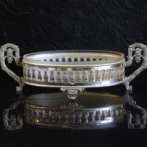 May include: A silver serving dish with a clear glass insert. The dish features ornate handles and a decorative balustrade design around the rim. It sits on a reflective surface, against a dark backdrop.