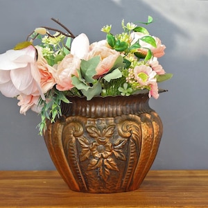 Op de afbeelding: Een bronzen decoratieve vaas gevuld met een boeket roze en witte bloemen en groen blad. De vaas heeft een sierlijk bloemmotief en staat op een houten oppervlak.