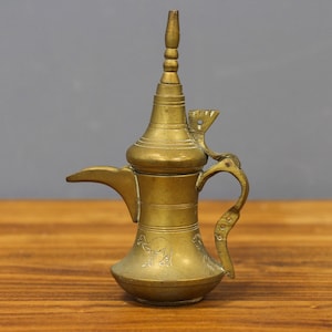 May include: A vintage brass coffee pot with a long spout, ornate handle, and decorative engravings. The pot has a tall, conical top and a bulbous base. The brass has a warm, aged patina, suggesting it is an antique or vintage item.