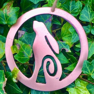 Puede incluir: Un adorno de metal de color cobre en forma de círculo con un recorte de una liebre o conejo en su interior. El adorno está colgado de una cuerda de hilo y está rodeado de hojas verdes.