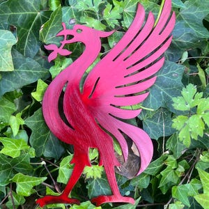 May include: A red metal cutout of the Liverpool Football Club Liver bird emblem, featuring the mythical bird standing with wings spread and holding a sprig of seaweed in its beak, showcasing a brushed metallic texture and sharp edges.