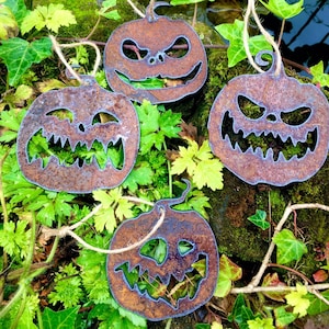 Może przedstawiać: Cztery rdzawe metalowe dekoracje dyni na Halloween ze straszną twarzą. Dynie wiszą na sznurku i są otoczone zielonym listowiem.