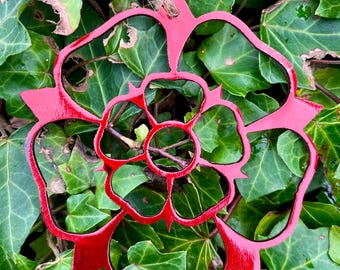 May include: Red metal rose ornament with a detailed design, hanging from a twine string. The ornament is on a bed of green leaves.
