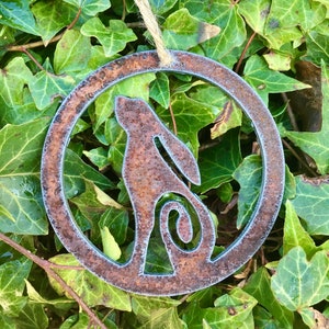 May include: A rusty metal ornament shaped like a circle with a cutout of a stylized rabbit inside. The ornament is hanging from a twine string.