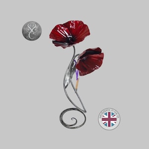 May include: A metal sculpture featuring two deep red poppy flowers with glossy petals attached to curved, silver-toned stems that spiral into a circular base. The sculpture includes a small circular logo with the text Handmade in the UK.
