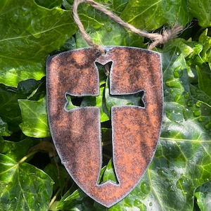May include: A shield-shaped metal ornament with a cross design cut out. The metal has a distressed, brown finish, and a natural rope for hanging. It is set against a background of lush, green ivy.
