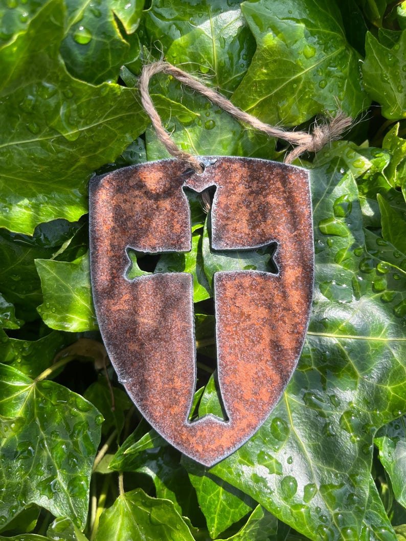 Może przedstawiać: Rustykalna, metalowa ozdoba w kształcie tarczy z wyciętym krzyżem. Metal ma zwietrzałe, brązowe wykończenie i naturalny sznurek do zawieszenia. Ozdoba jest umieszczona na tle żywego, zielonego bluszczu.