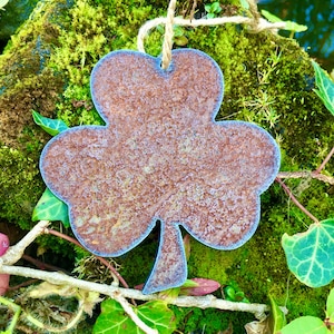 Könnte beinhalten: Ein rostiger Metallshamrock-Anhänger mit rustikalem, gealtertem Aussehen. Der Kleeblatt hängt an einer Bindfadenkordel und ist von grünem Moos und Efeu umgeben.