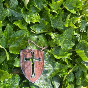 Może przedstawiać: Brązowa ozdoba w kształcie tarczy z wyciętym wzorem krzyża, zawieszona na sznurku. Ozdoba umieszczona jest na tle żywych, zielonych liści bluszczu, tworząc naturalną, ziemistą estetykę. Krzyż jest dominującym elementem.