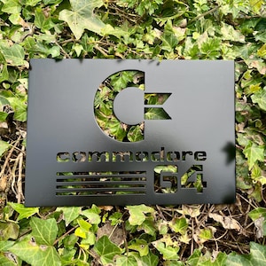 May include: Black metal sign with a white Commodore 64 logo. The logo features a stylised "C" with a diagonal line through it. The text "Commodore 64" is below the logo.