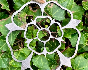 May include: A silver metal rose ornament with a detailed design. The ornament is shaped like a rose with a central circle and multiple petals.