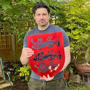 May include: A red shield featuring a cutout design of three black lions stacked vertically. The shield has a smooth, flat surface with a curved top edge and a pointed bottom, displaying a heraldic-style motif with sharp, clean edges.