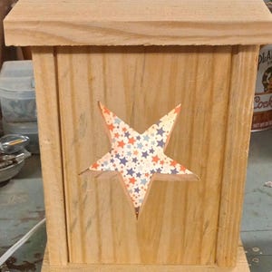 May include: A wooden box with a star-shaped cutout on the front. The star is filled with a patterned paper featuring red, white, and blue stars. The box is made of light-colored wood and has a simple, rectangular design.