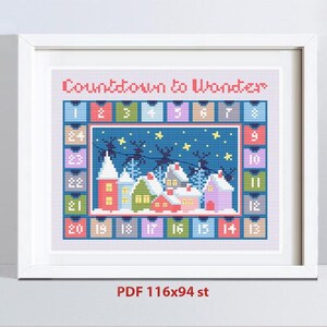 May include: Framed cross-stitch art with a winter village scene. The artwork features colorful houses, a starry night sky, and the text "Countdown to Wonder." The frame is white.