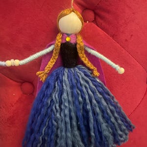 May include: A handmade felt doll with brown hair, a purple cape, and a blue and gray yarn skirt. The doll has a wooden head and black beads for eyes.