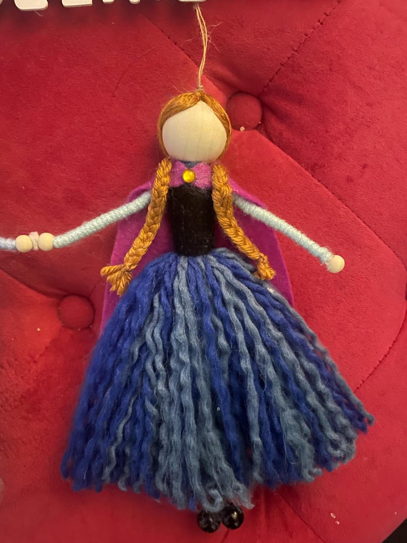 May include: A handmade felt doll with brown hair, a purple cape, and a blue and gray yarn skirt. The doll has a wooden head and black beads for eyes.