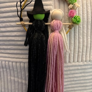 以下が含まれることがあります： 2つの糸人形を特徴とする手作りのハロウィーンの装飾。1つの人形は黒い帽子と黒い糸の髪を持つ魔女で、もう1つはピンクと白の糸の髪を持つ人形です。人形は花で飾られた金のフープに取り付けられています。