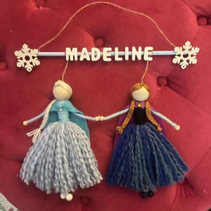 May include: A personalized mobile with the name "Madeline" in white letters on a blue bar. Two felt dolls, one in blue and one in purple, are hanging from the bar. Each doll has a snowflake attached to its head.