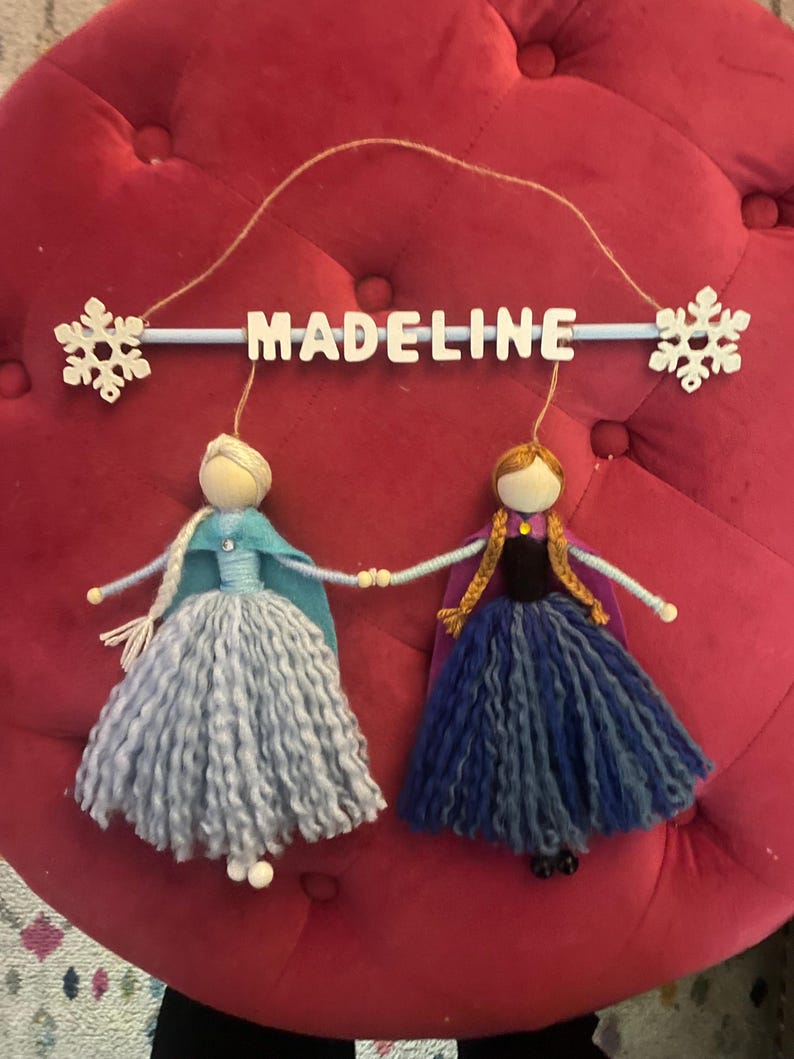 May include: A personalized mobile with the name "Madeline" in white letters on a blue bar. Two felt dolls, one in blue and one in purple, are hanging from the bar. Each doll has a snowflake attached to its head.